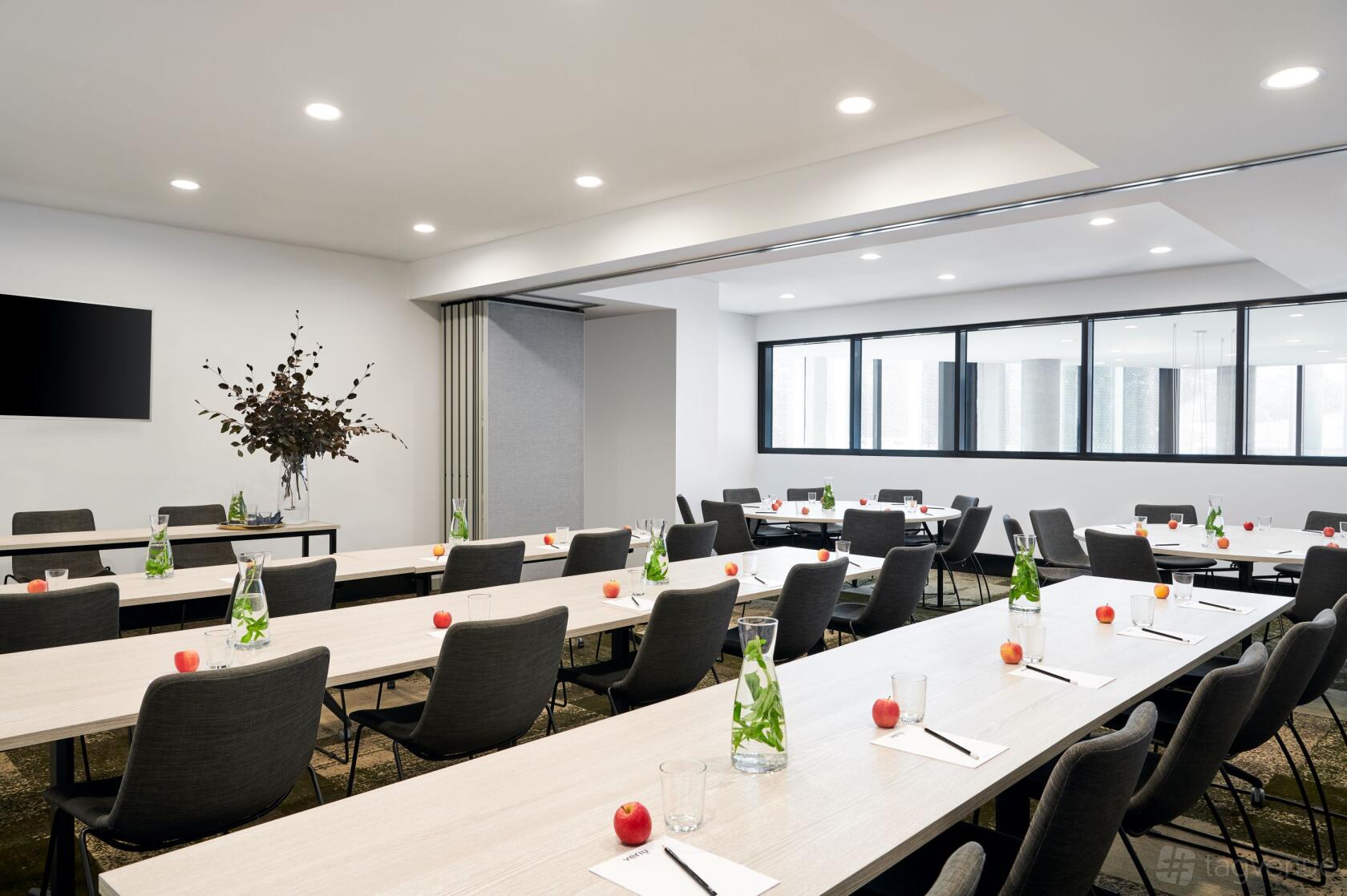 A hotel event room with rows of tables and chairs, bottled water, apples, and notepads at Veriu Green Square.