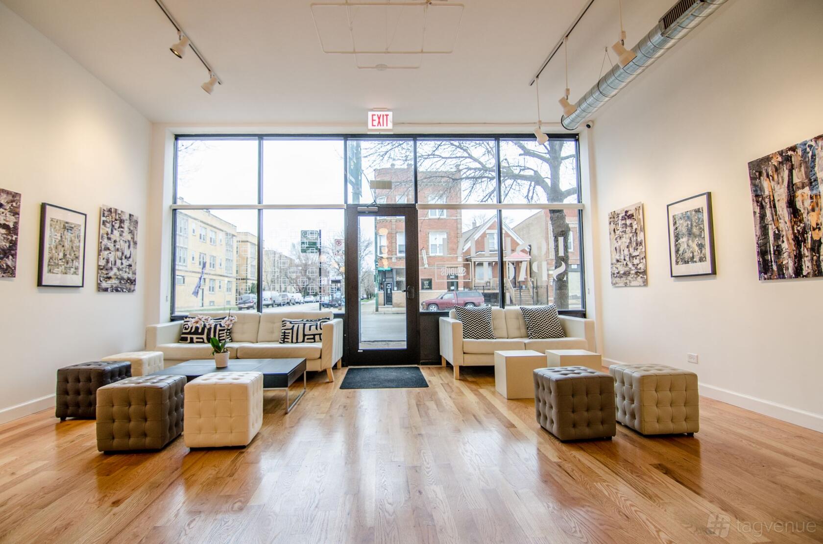 A gallery with floor-to-ceiling windows, wooden floors, modern seating, and artwork on white walls at Positive Space Studios.