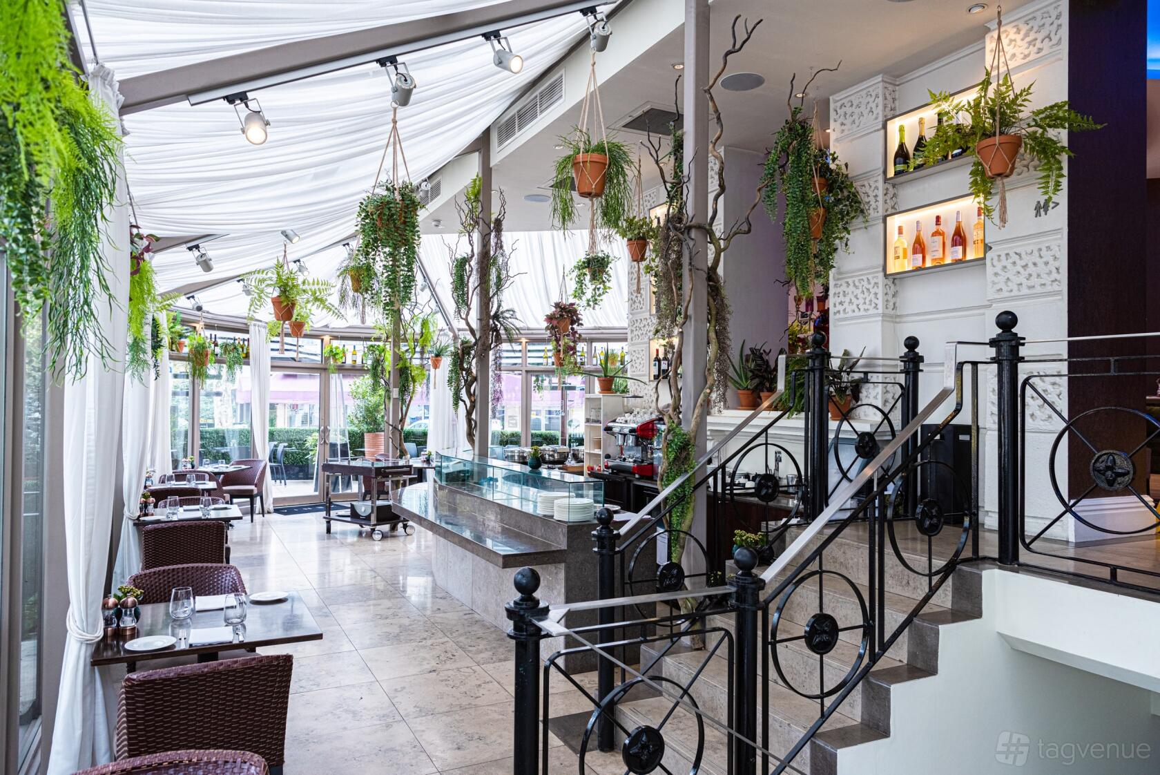 A conservatory restaurant with glass walls, draped white ceilings, potted plants, and set dining tables at La Terrazza.