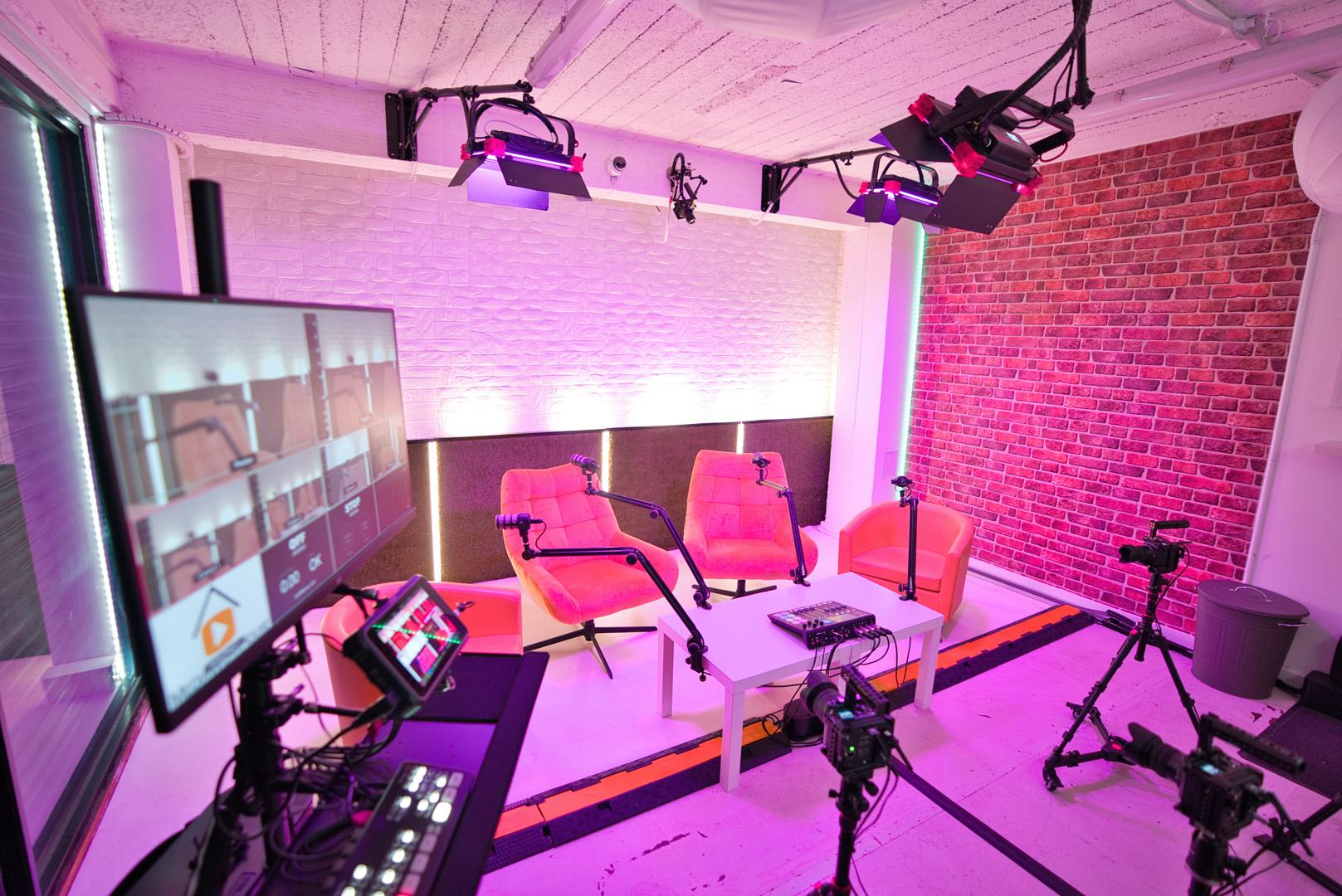 A podcast studio with pink armchairs, exposed brick walls, microphones, cameras, and studio lighting at MediaWorkHouse.