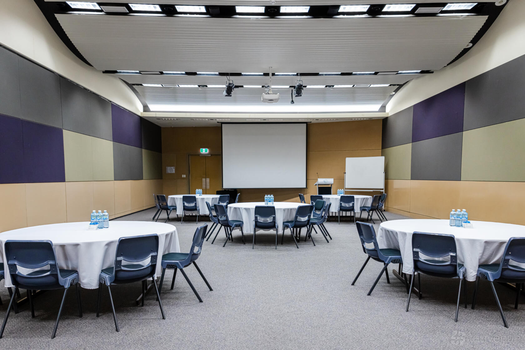 An auditorium with round tables covered in white linens, a projector screen, and colored acoustic wall panels at BTP Conference & Exhibition Centre.