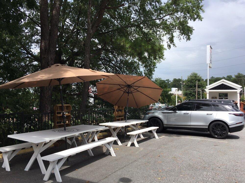 An outdoor restaurant area with white picnic tables, tan umbrellas, and surrounding trees at Cubanos ATL - SANDY SPRINGS TINY HOUSE.