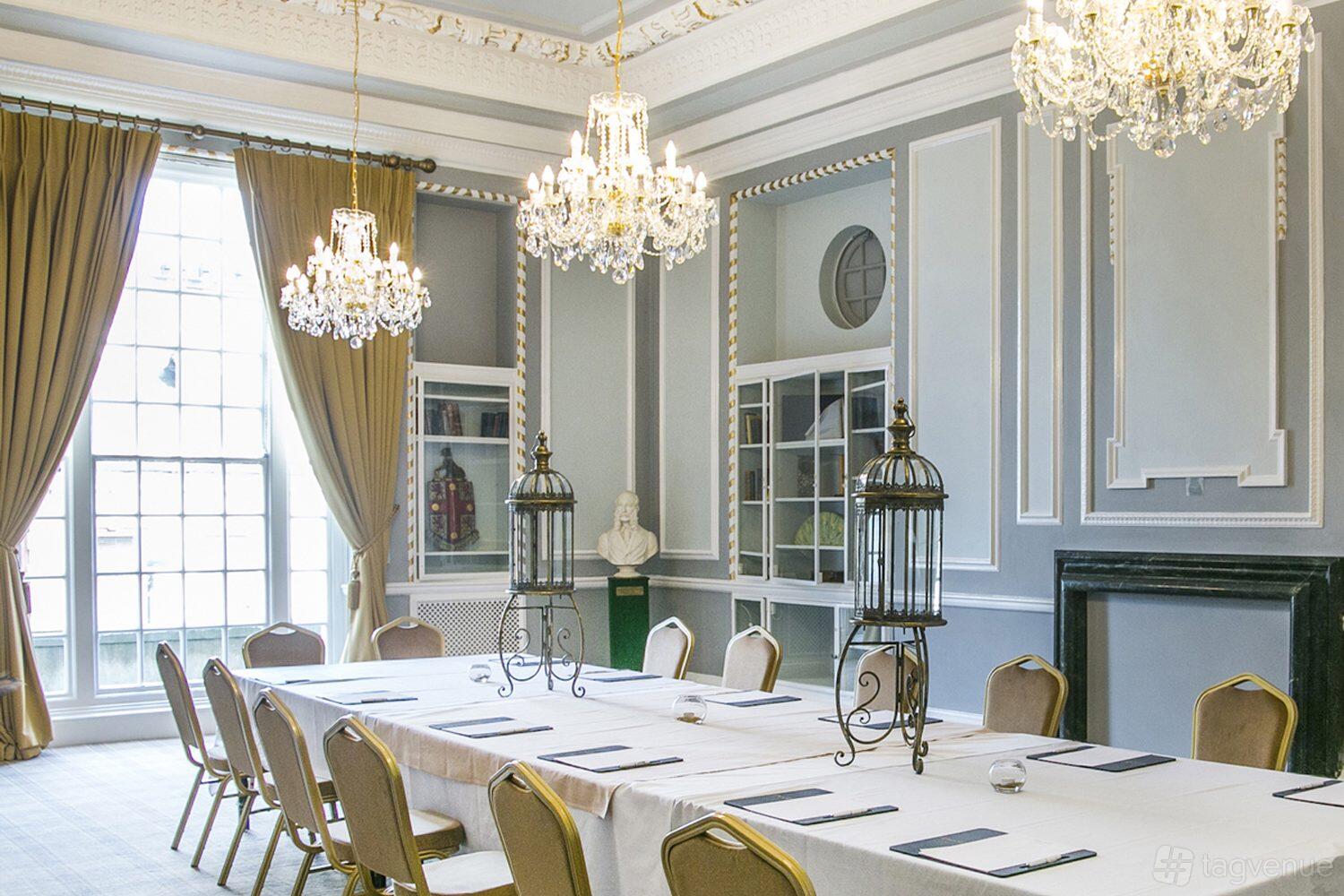 An event space with chandeliers, ornate molding, tall windows, and a long table with white linens at Manchester Hall.