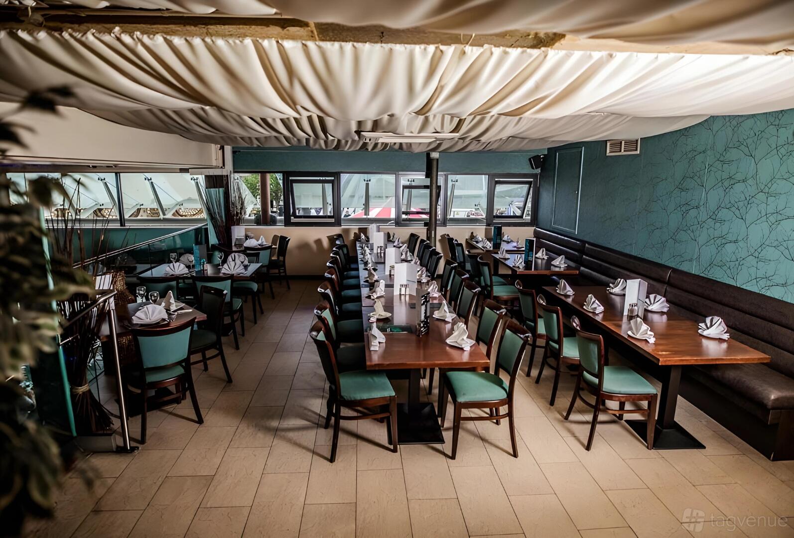 A restaurant with long wooden tables, teal chairs, napkin place settings, and draped ceiling decor at Tikka Flame.