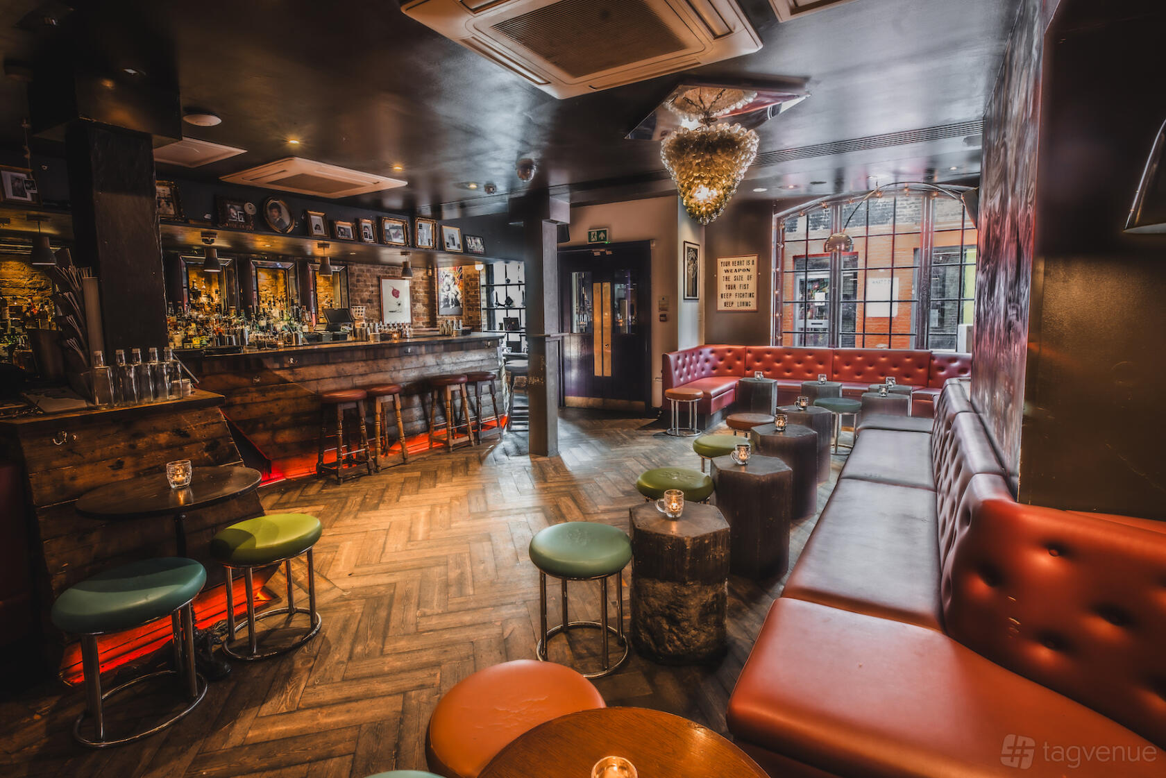 A cocktail bar with a wooden herringbone floor, red banquette seating, round stools, and a chandelier at Callooh Callay Shoreditch.