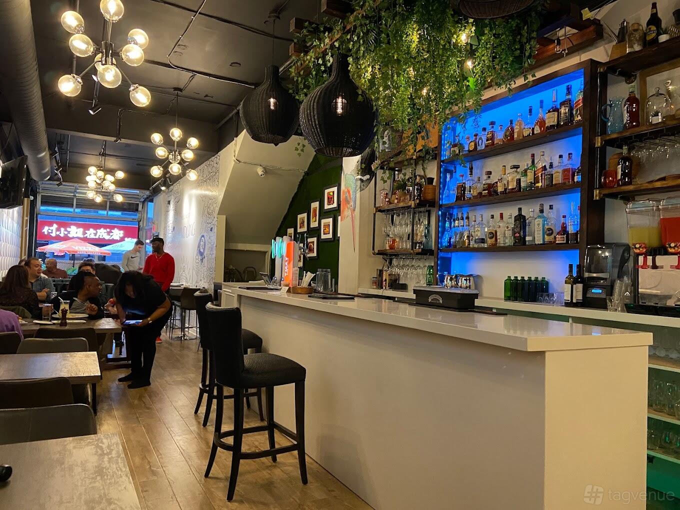 A restaurant with a white bar counter, illuminated blue shelving, hanging greenery, and modern pendant lights at The Diner's Corner.