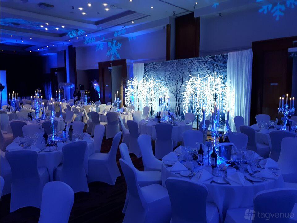 An event space in a hotel with round tables draped in white linens, candelabras, and blue winter-themed lighting at Chesford Grange.
