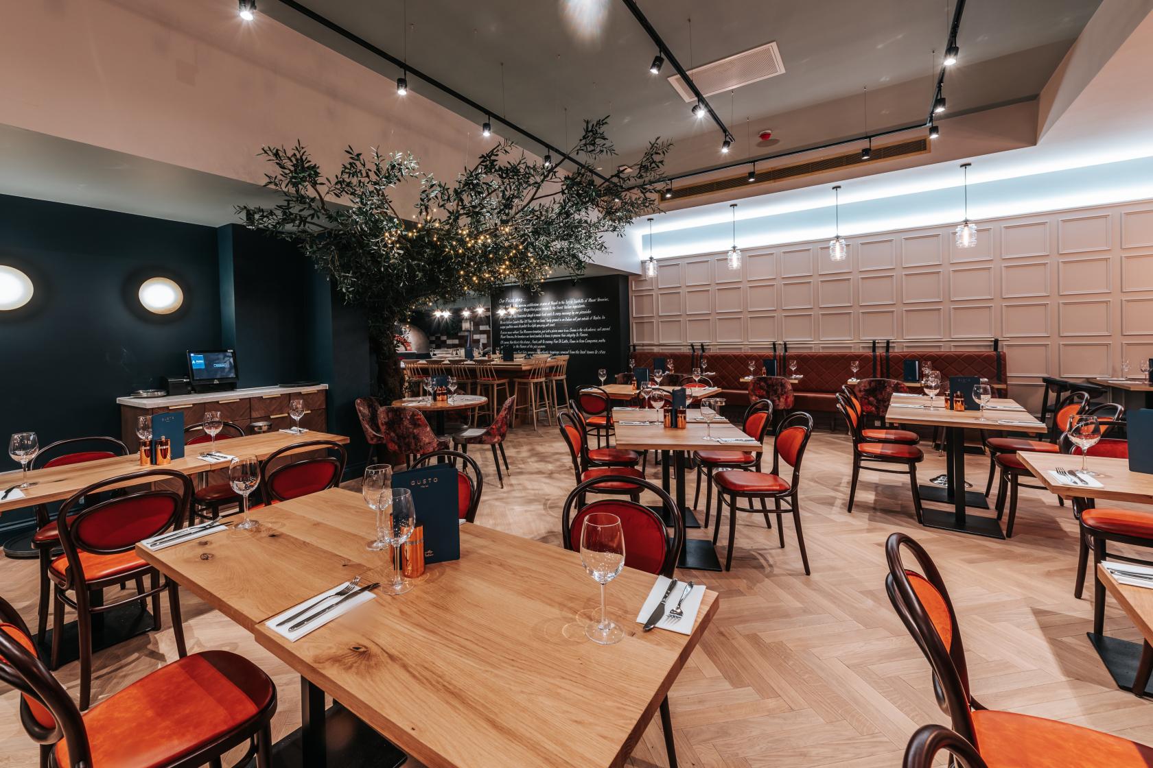 A restaurant with wooden tables, red chairs, parquet flooring, and an artificial tree at Gusto Italian Nottingham.