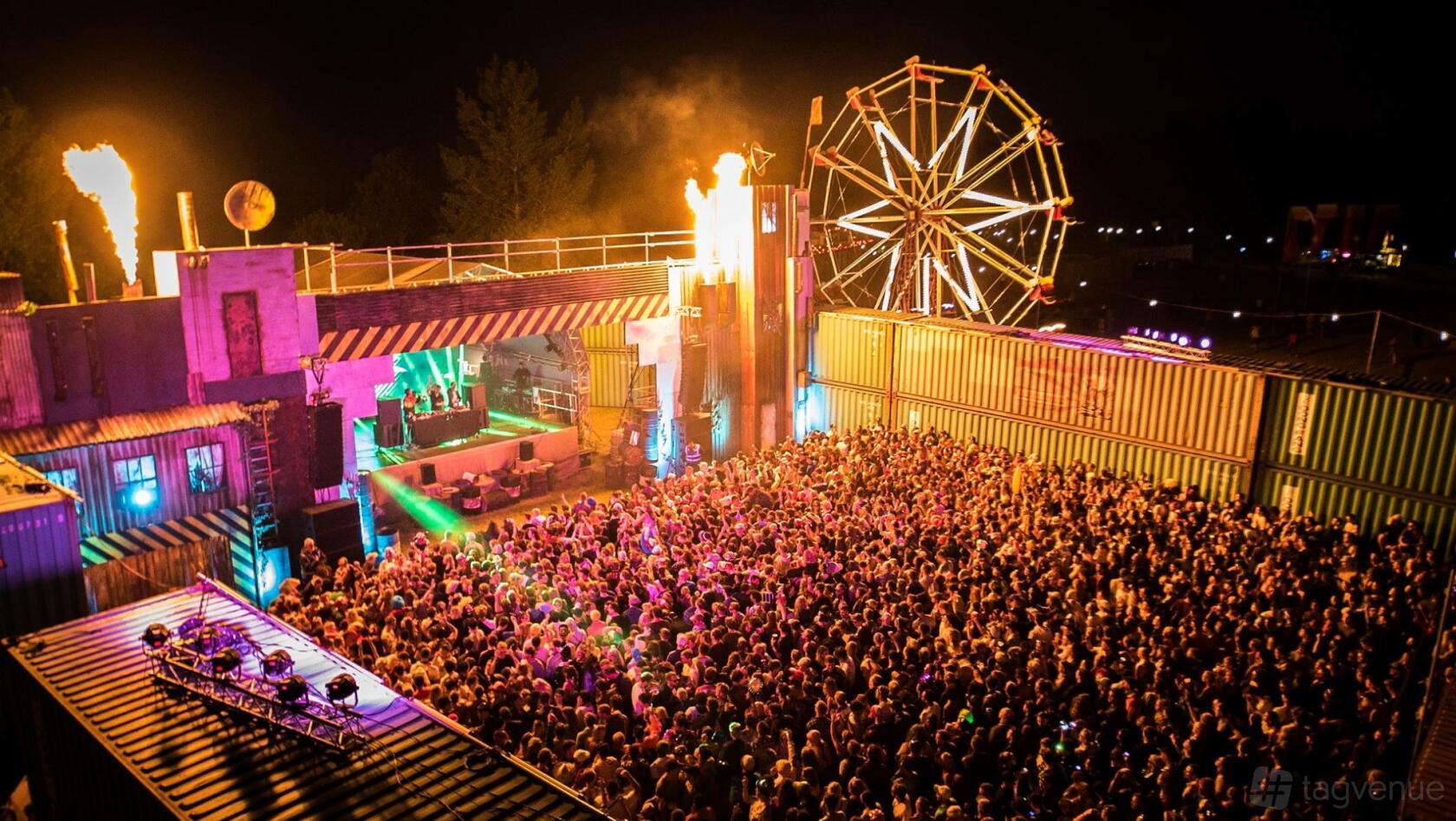 An open air venue with a large festival crowd, stage lighting, and a Ferris wheel at Bygrave Woods.