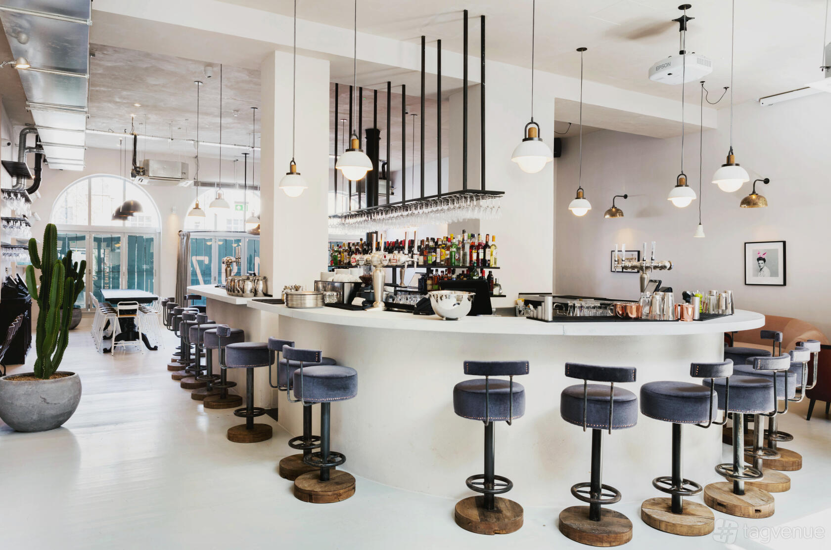 A bar with a curved white counter, velvet barstools, pendant lighting, and large windows at No.197 Chiswick Fire Station.