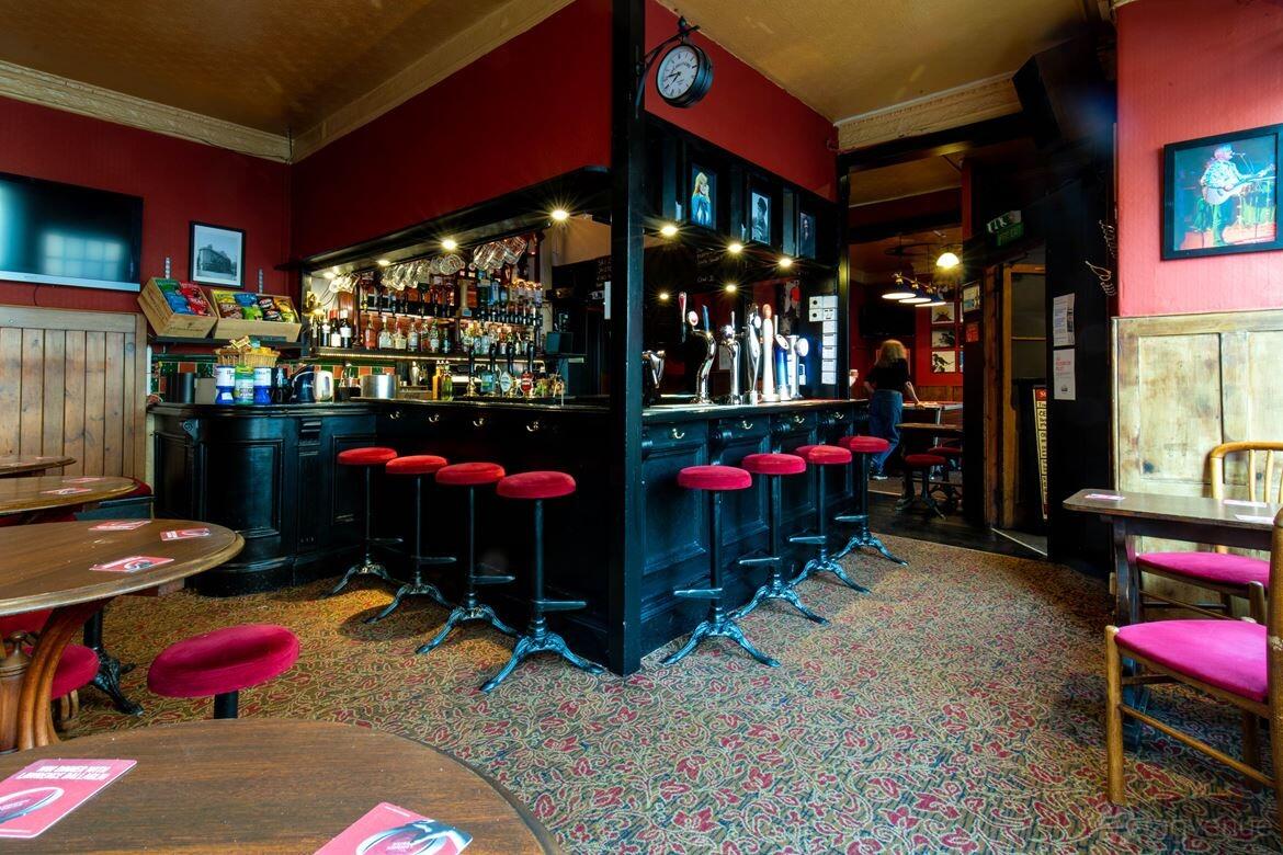 A pub with a carpeted floor, red upholstered barstools, wooden tables, and a corner bar at St James Wine Vaults.