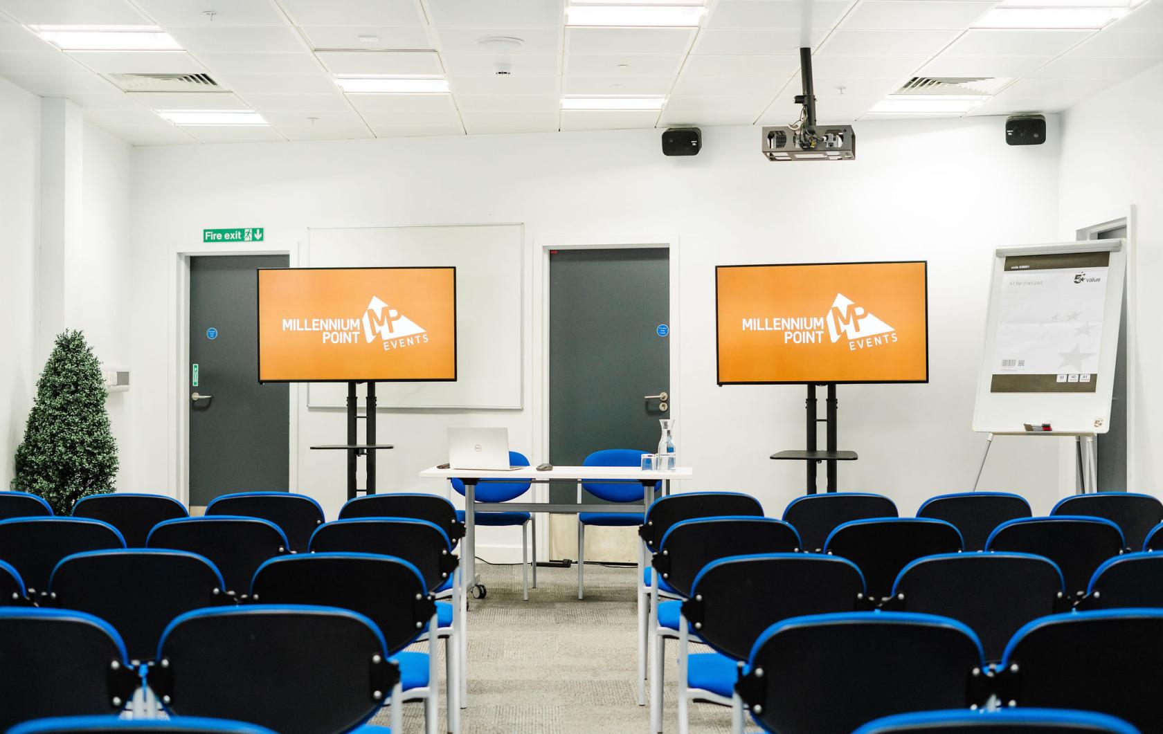 A conference centre with rows of blue chairs, dual presentation screens, and a presenter table at Millennium Point.