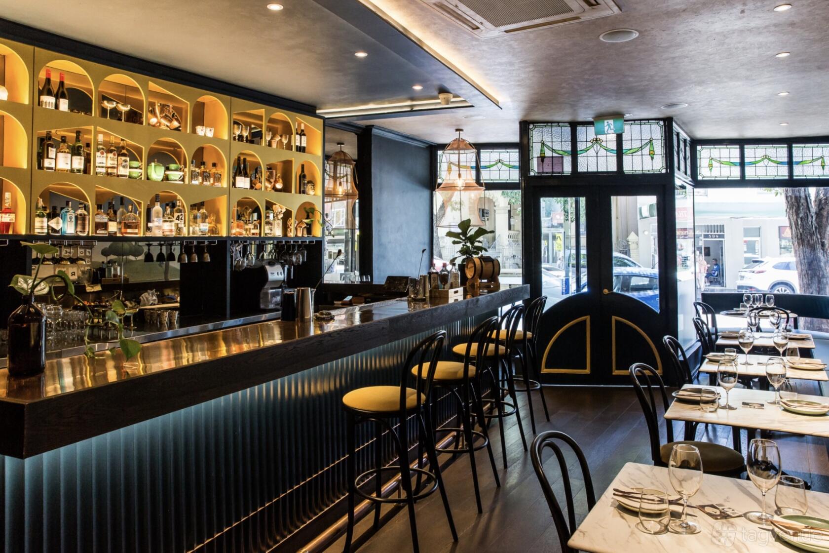 A restaurant with a curved bar, backlit shelves displaying bottles, and tables set with glassware at Foreign Return Indian Restaurant.
