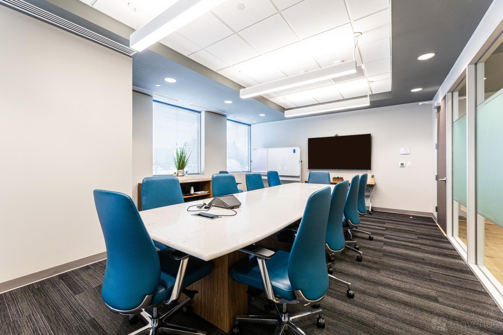 A boardroom with a long white conference table, blue ergonomic chairs, large windows, and a wall-mounted screen at MedtoMarket Event and Conferencing Center.
