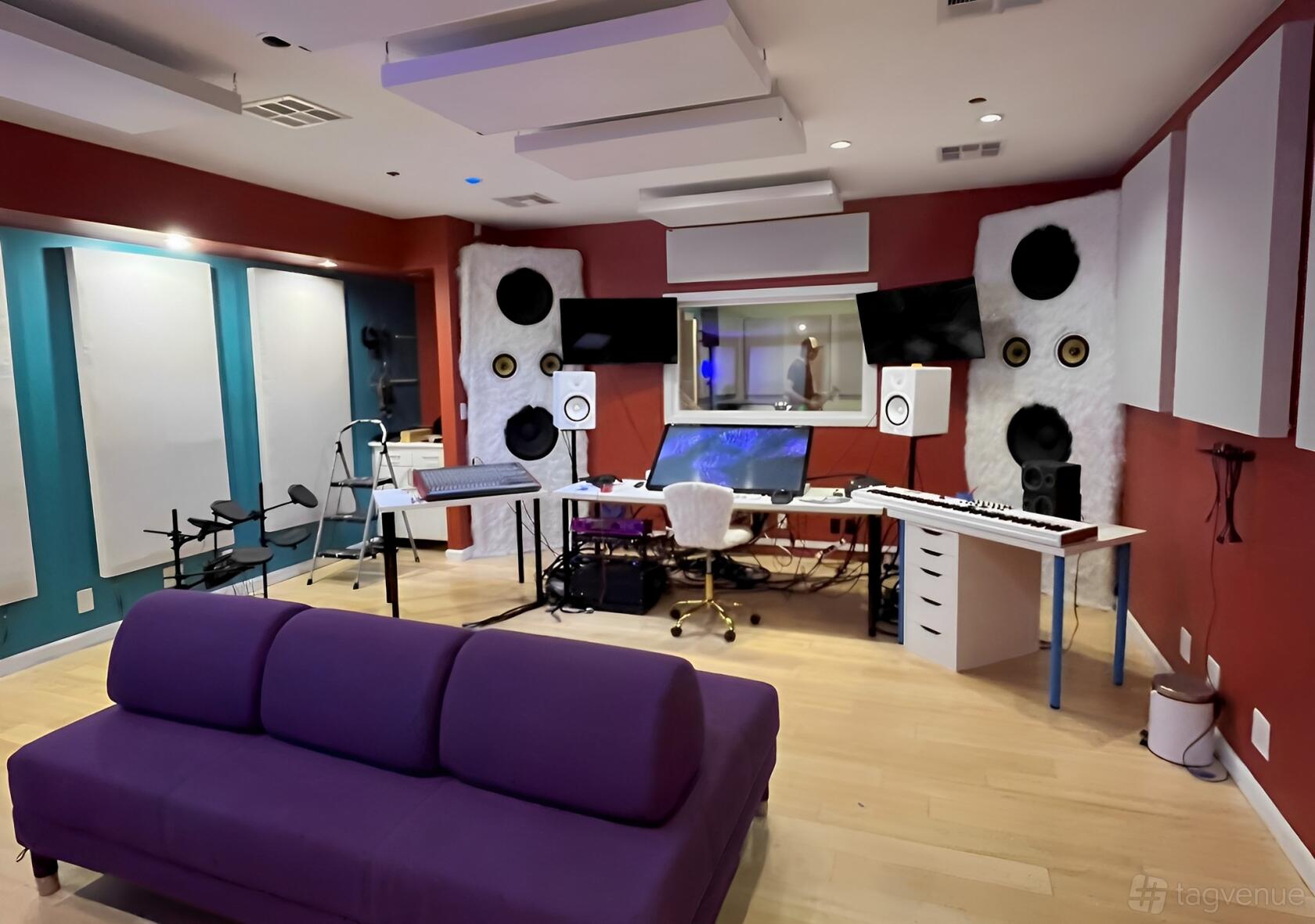A recording studio with acoustic panels, mixing desk, dual monitors, and a purple couch at The Music Company.