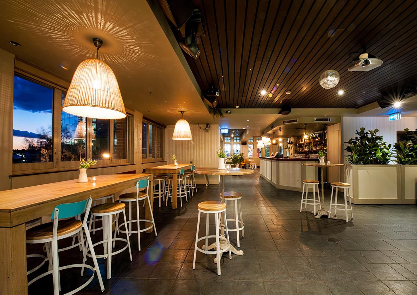 A hotel bar with wooden tables, bar stools, pendant lighting, and large windows at Shelbourne Hotel's LinQ Bar.