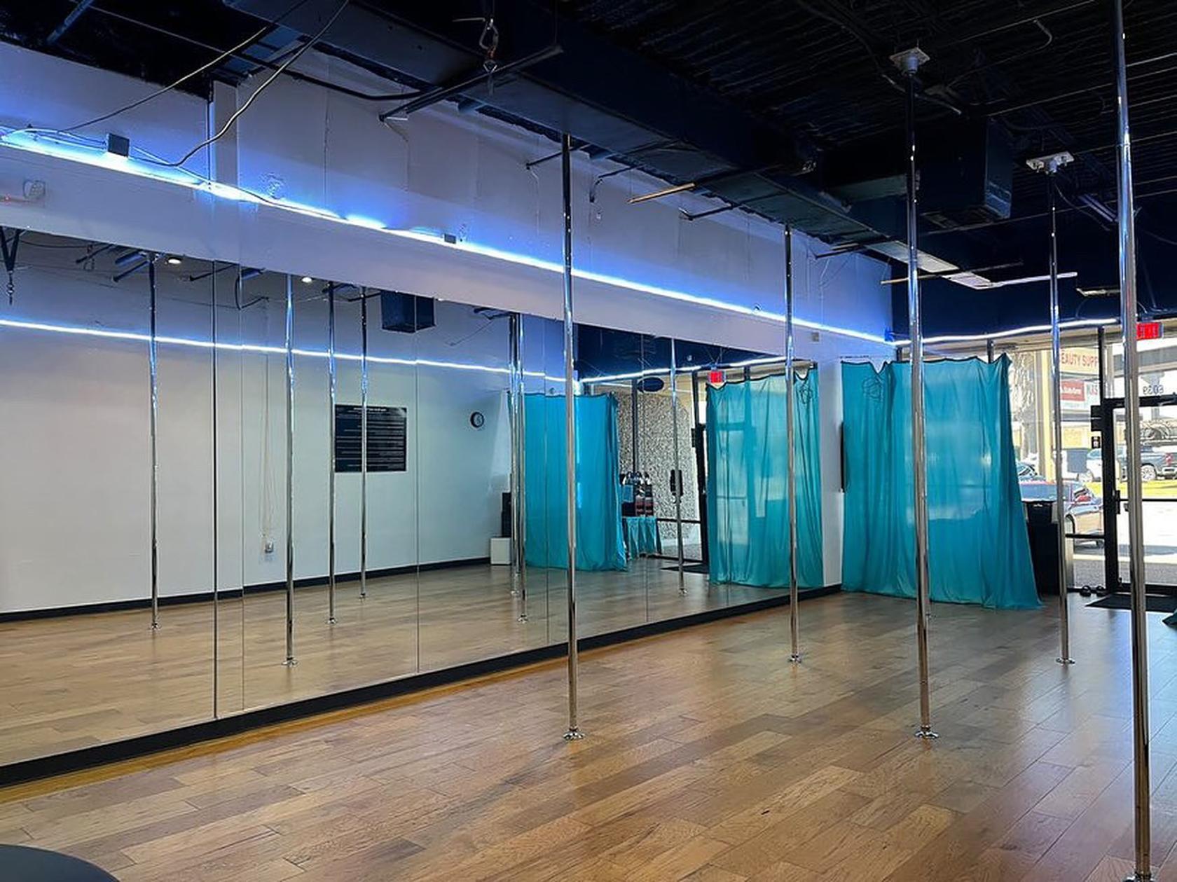 A dance studio with mirrored walls, pole installations, wooden floors, and blue curtains at Brass Ovaries.