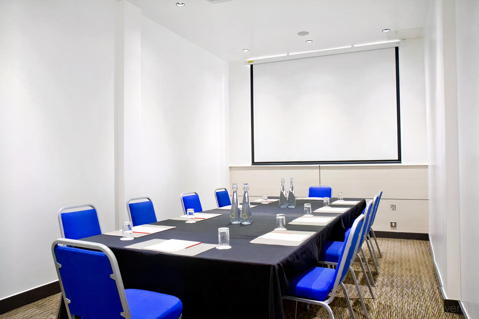 A hotel meeting room with a long table, blue chairs, notepads, water bottles, and a projector screen at The Wesley Euston Hotel.
