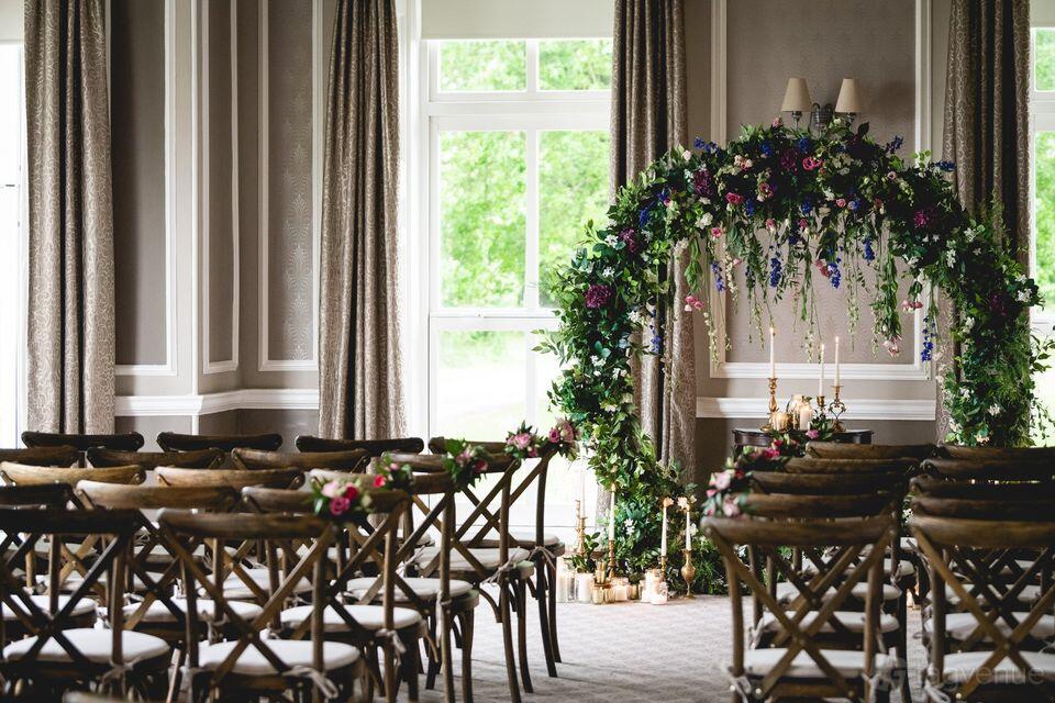 A hall with wooden chairs arranged in rows, large windows, and a floral archway at Hothorpe Hall.