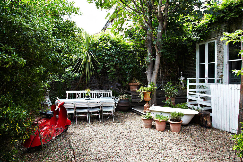 An outdoor event venue with gravel ground, long dining tables, potted plants, and a vintage bathtub at The Roost.