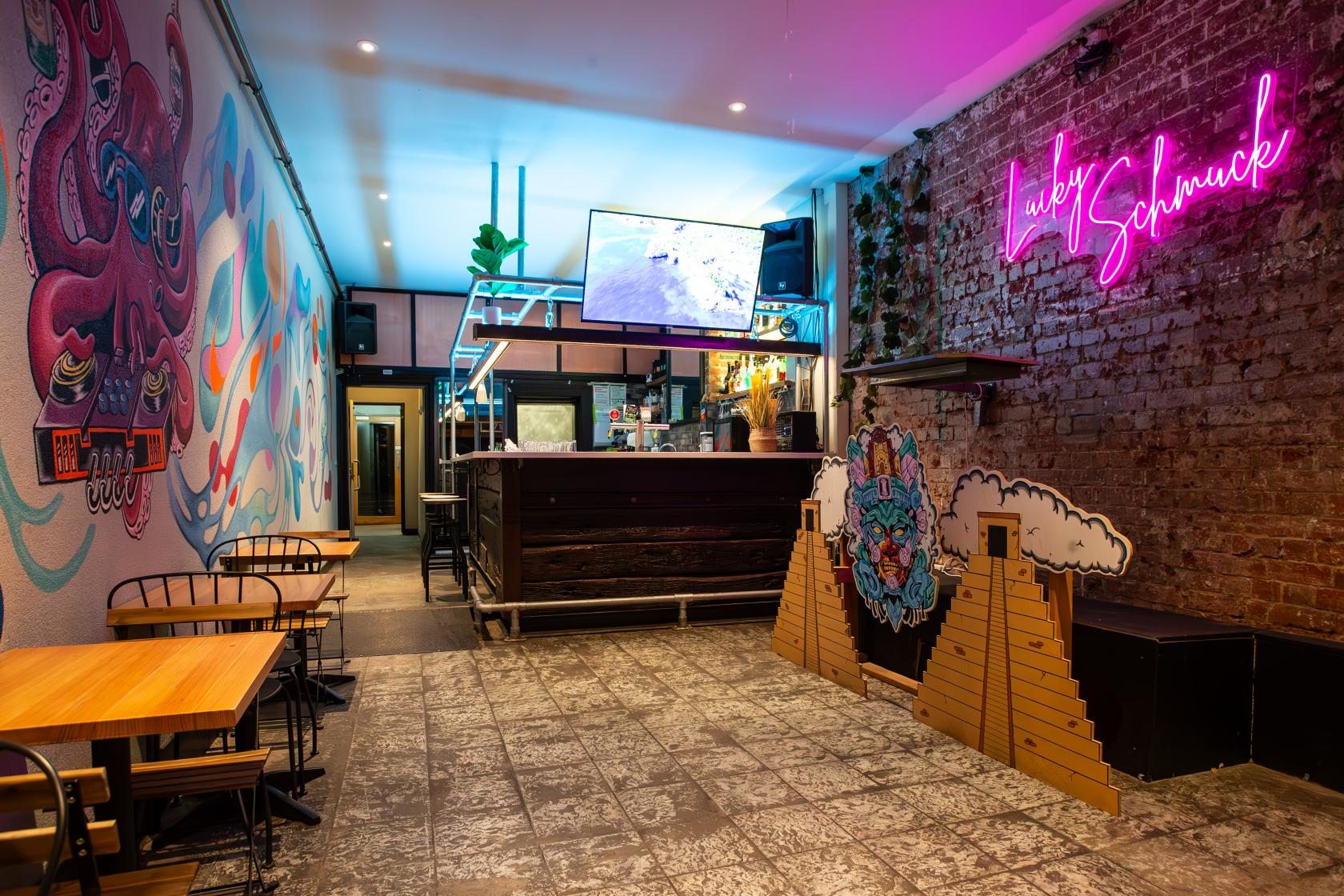 A bar with exposed brick walls, neon signage, colorful murals, and wooden tables at Lucky Schmuck.