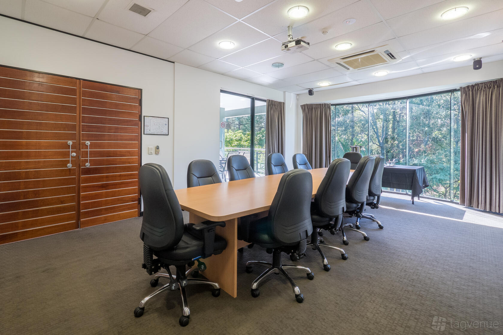 A boardroom with a long wooden table, black office chairs, floor-to-ceiling windows, and a projector at M1 Business Centre.