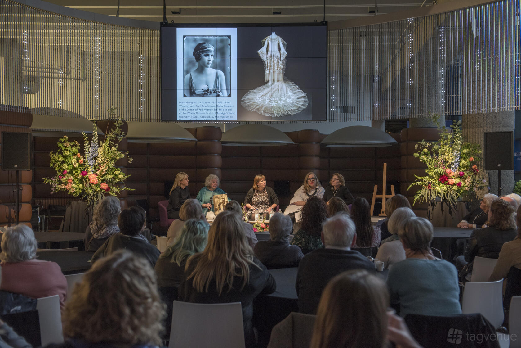 A museum hall with a panel discussion setup, large floral arrangements, and a screen displaying historical images at London Museum Spaces.