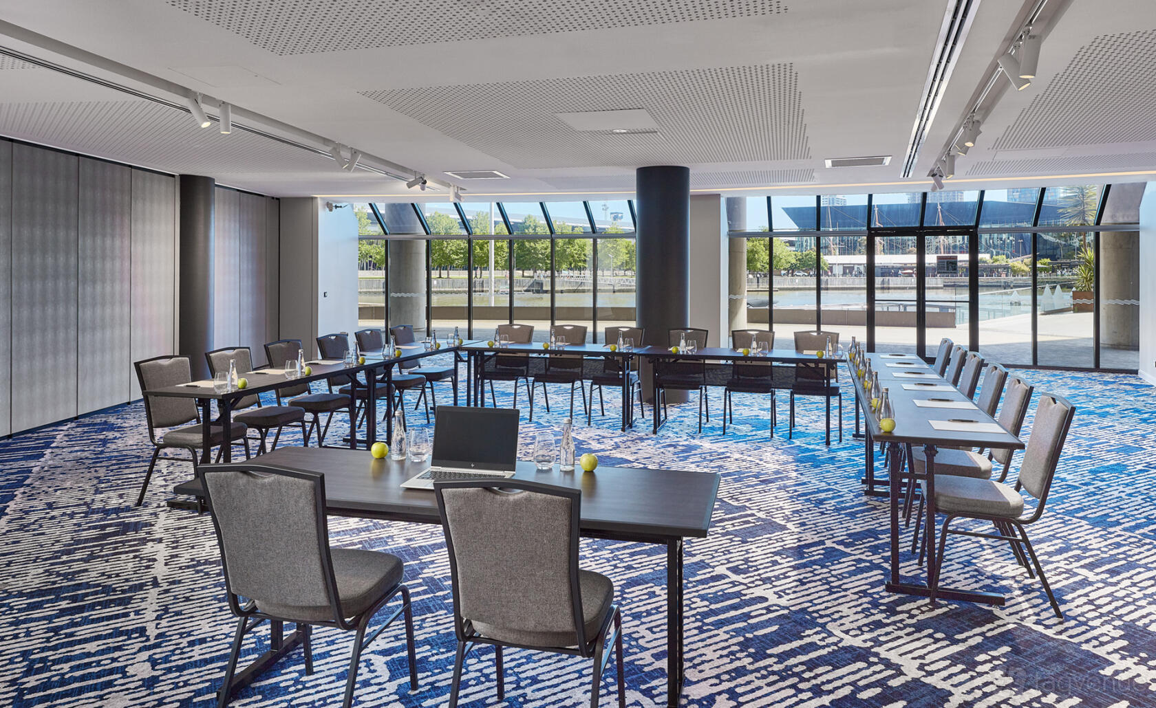 A meeting room with floor-to-ceiling windows, U-shaped tables, and patterned carpet at Pearl Riverfront.