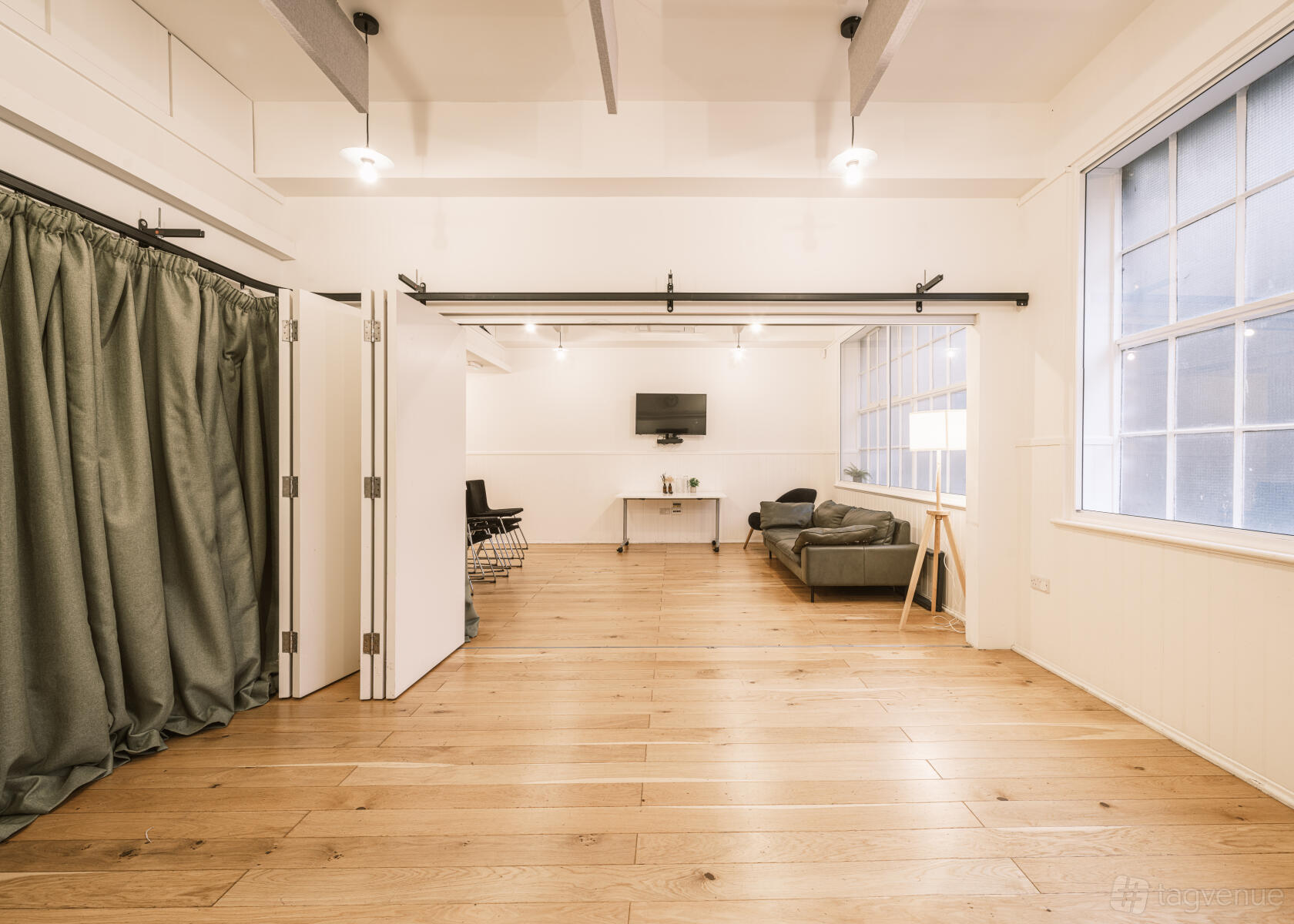A meeting room with light wood floors, large industrial windows, a sofa, and wall-mounted TV at King's House.