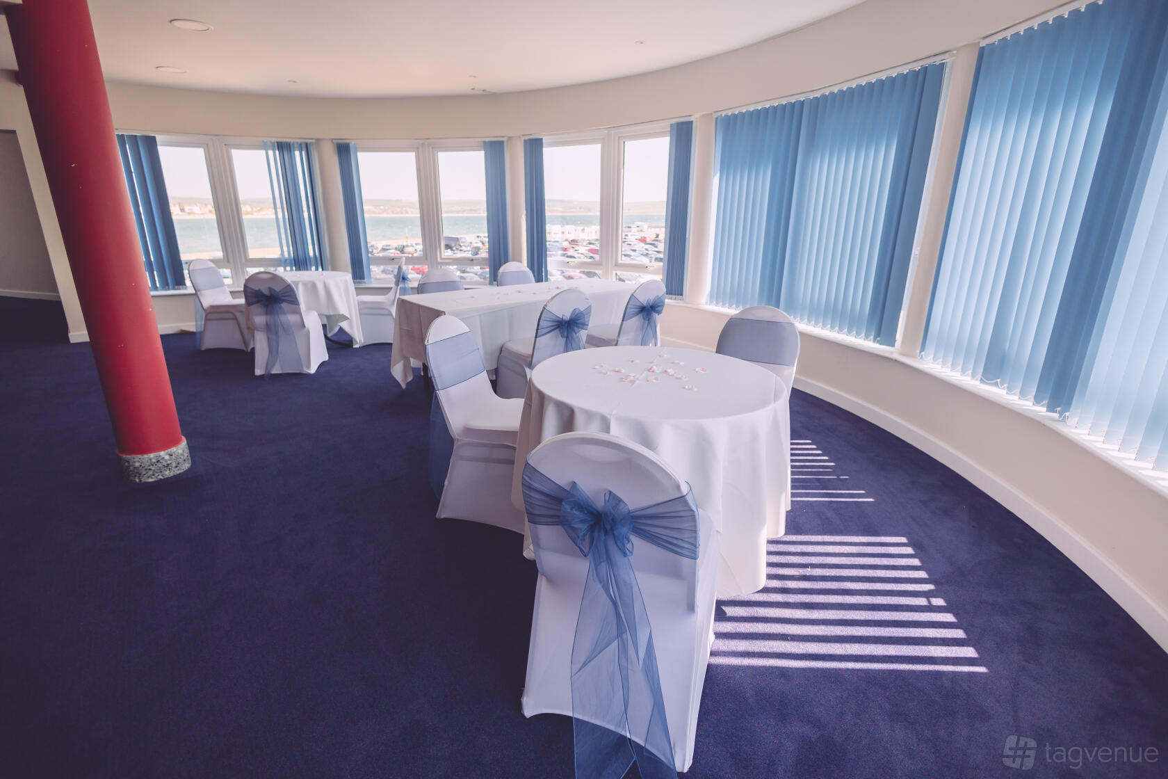 An event venue with round tables draped in white linens and blue sashed chairs by large windows at Weymouth Pavilion.
