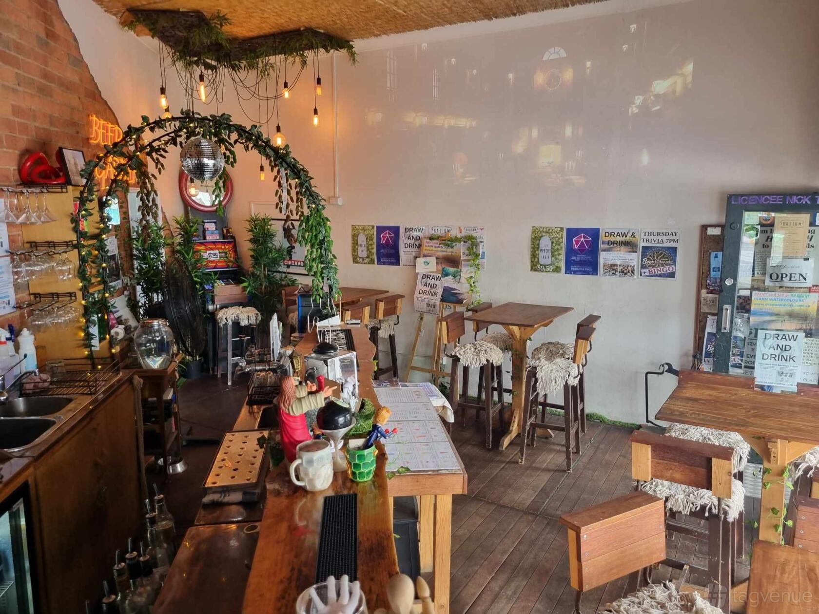 An indoor bar with hanging lights, wooden tables, a leafy arch over the counter, and posters on the wall at Radium Bar.