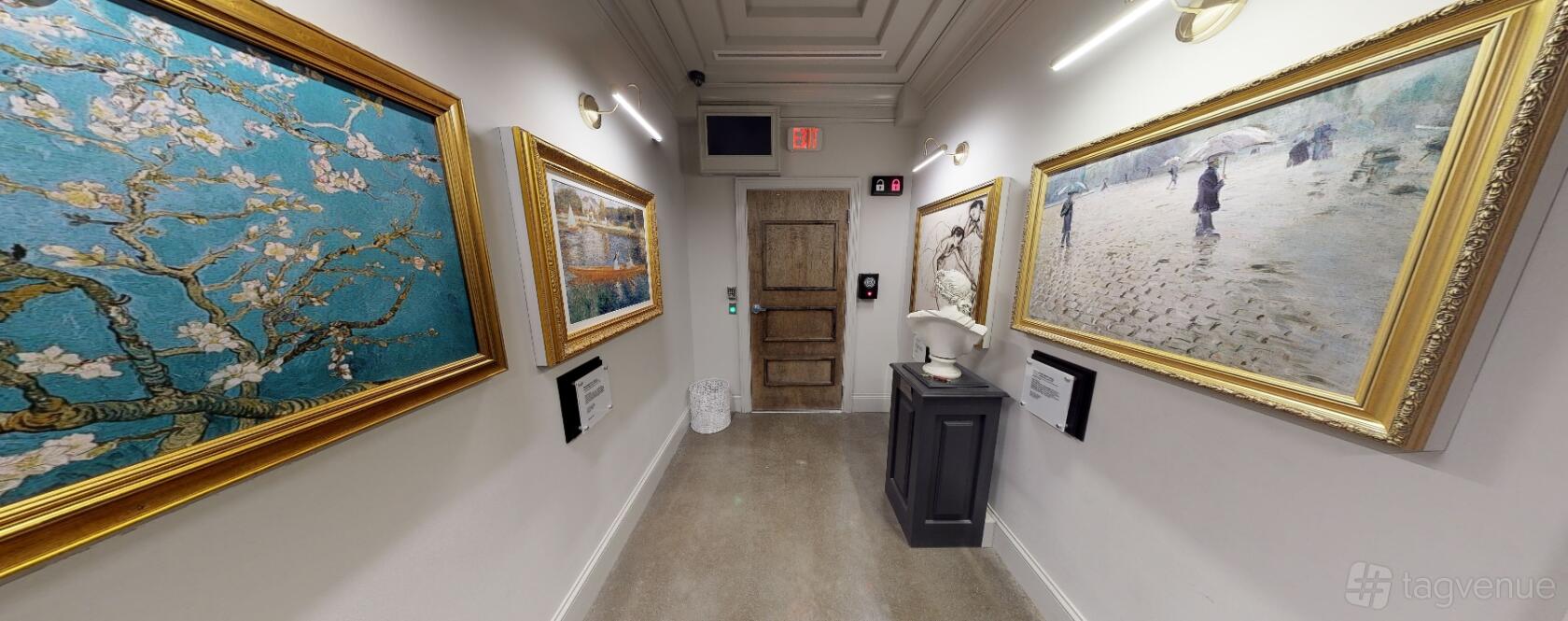 An event venue hallway with framed artwork, wall lighting, and a sculpture pedestal at The Escape Game Houston- The Galleria.