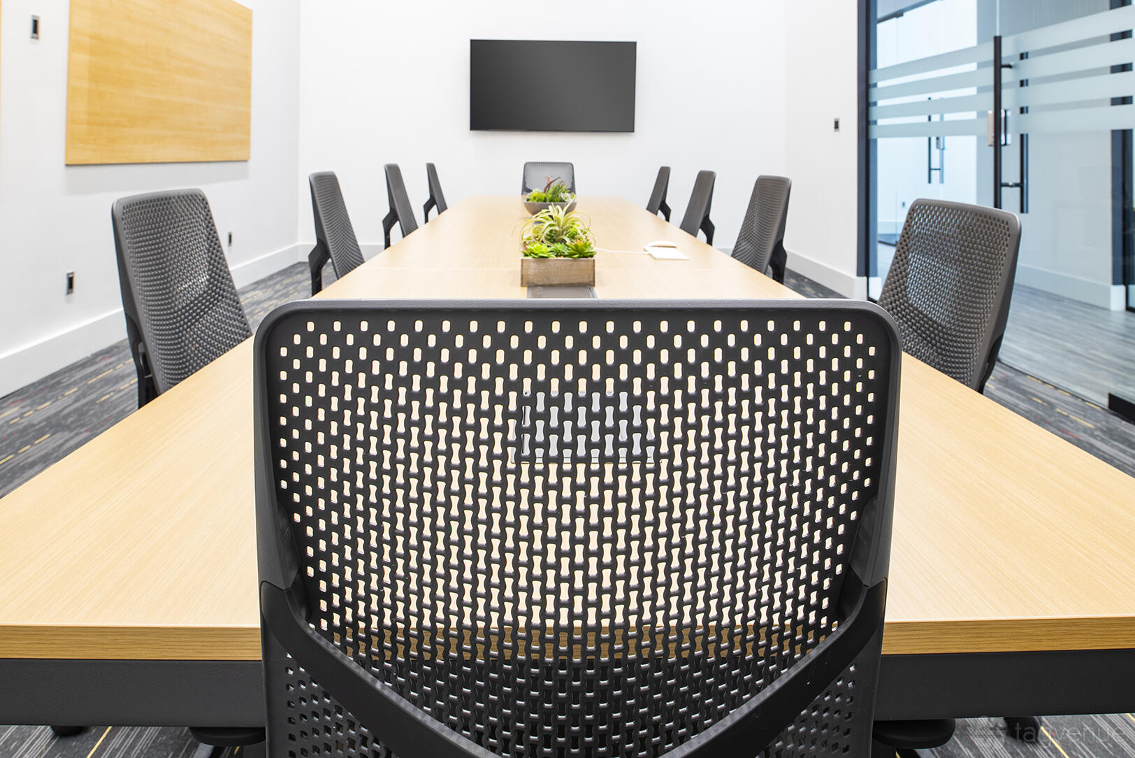 A meeting room with a long wooden conference table, mesh chairs, a wall-mounted screen, and a small plant at Venture X Heartland.