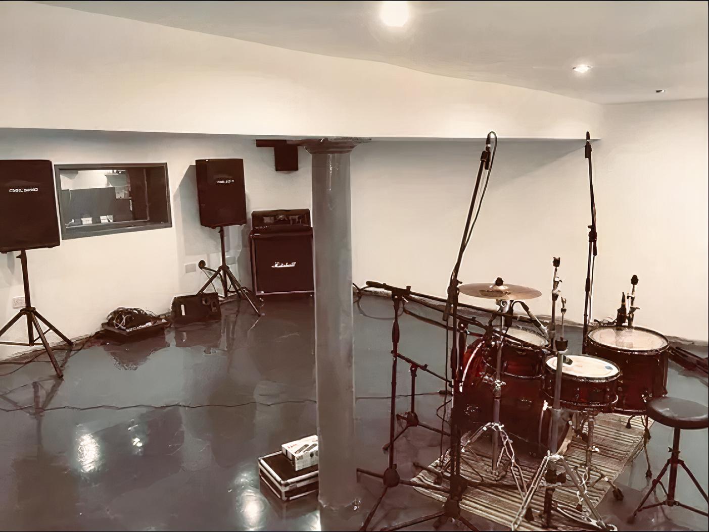 A rehearsal studio with drum kit, microphone stands, amplifiers, and a viewing window at Rain City Studios.