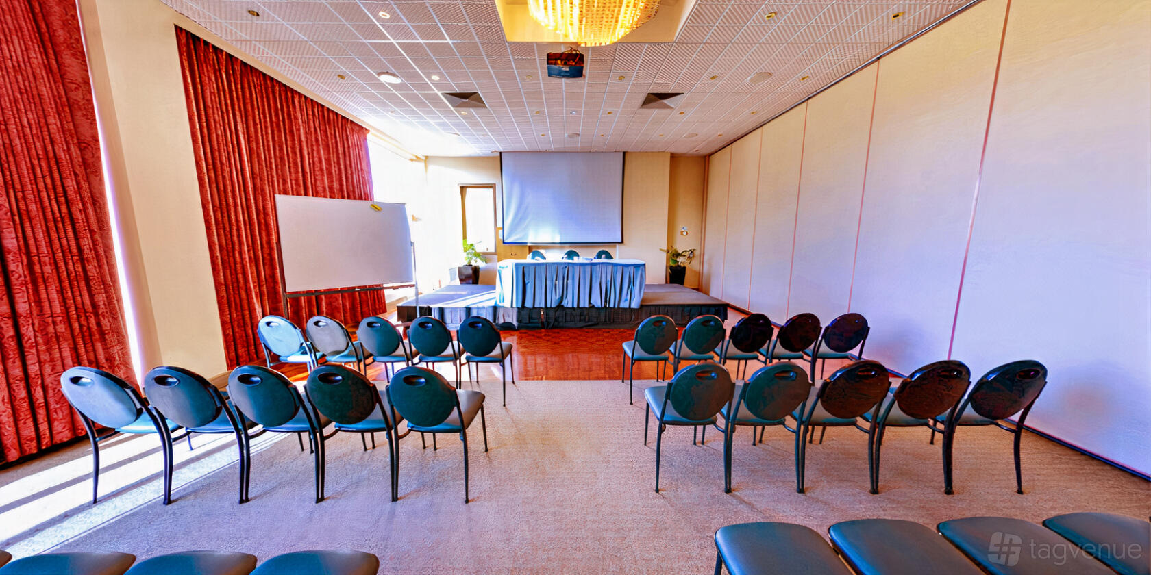 A meeting room with theater-style chairs, a stage with a table and microphones, and a projector screen at Best Western Airport Motel & Convention.