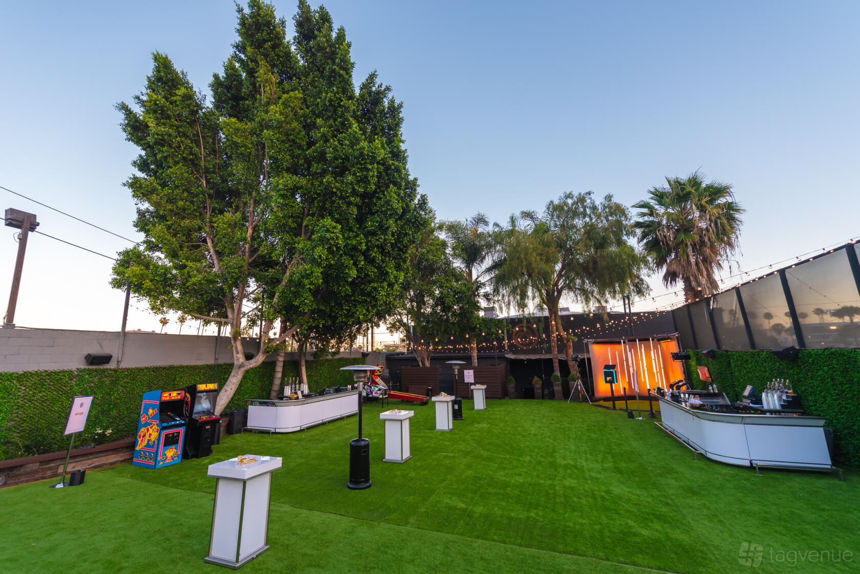 An outdoor nightclub patio with artificial grass, string lights, arcade games, and cocktail tables at Academy LA.