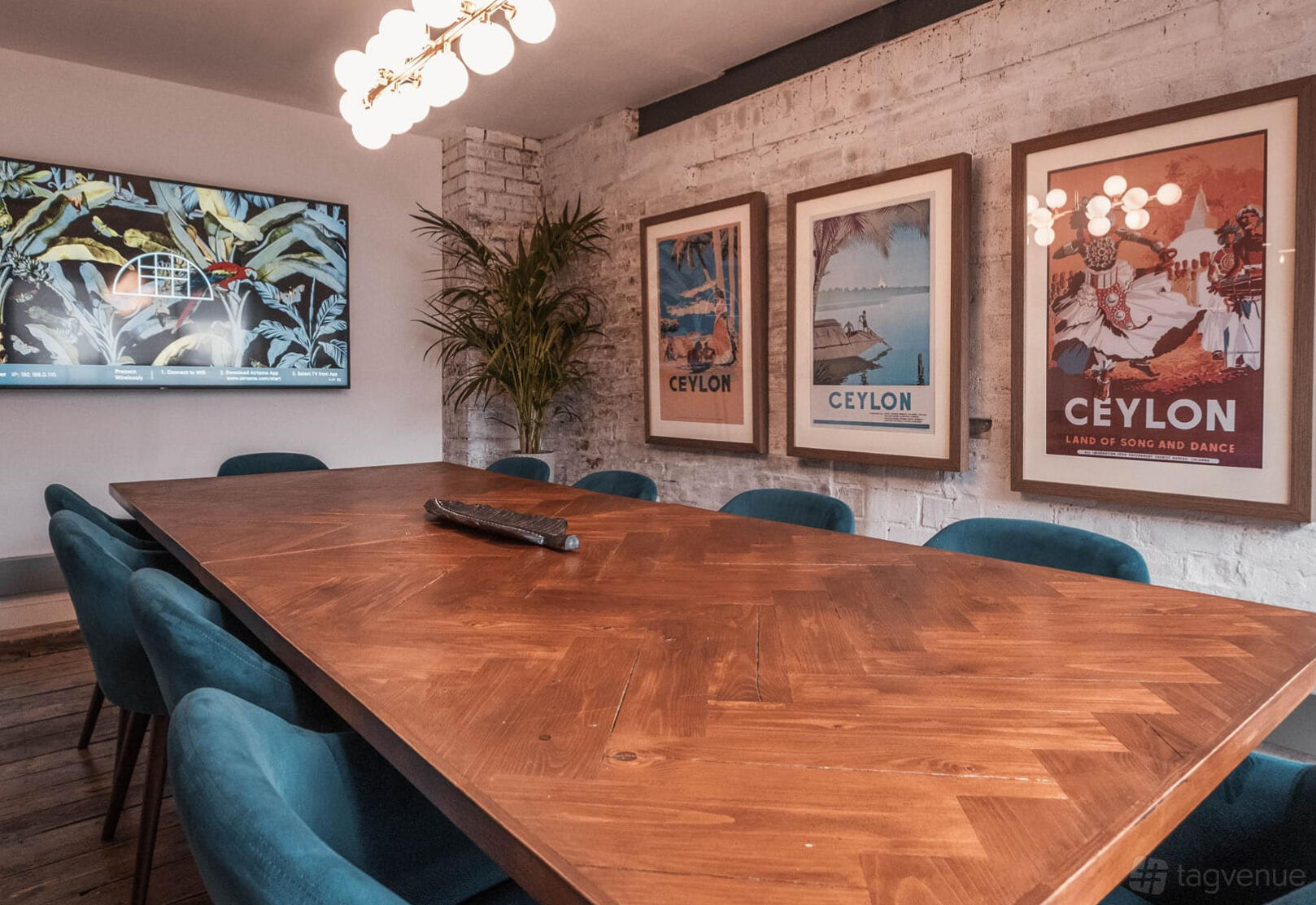 A meeting room with a herringbone wooden table, blue chairs, wall art, and a TV screen at Beehive Lofts.