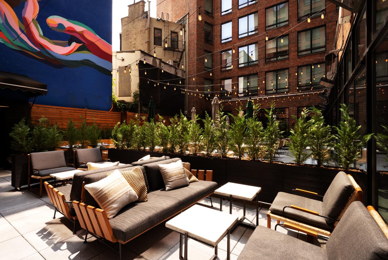 An outdoor hotel bar with lounge seating, potted greenery, string lights, and urban mural at Arlo Soho.