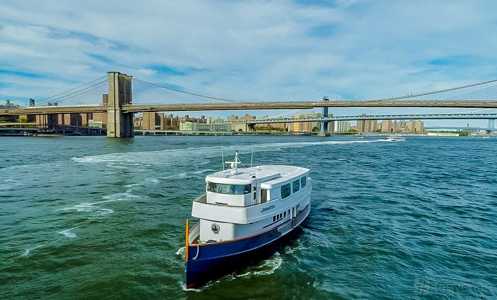 A boat with a white upper deck and blue hull cruising on the river near a suspension bridge at Chelsea Piers Marina.