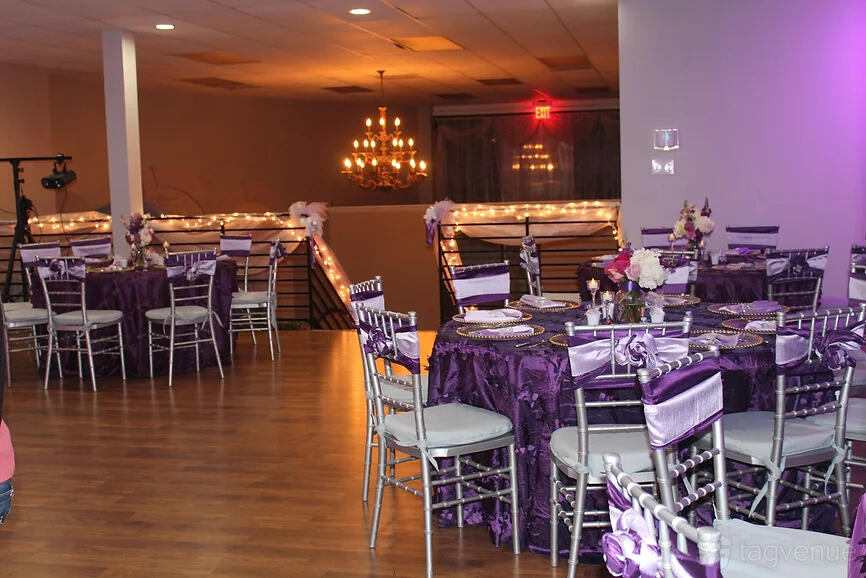 An event hall with round tables dressed in purple linens, silver chairs, wood flooring, and a chandelier at Venue At Memorial.