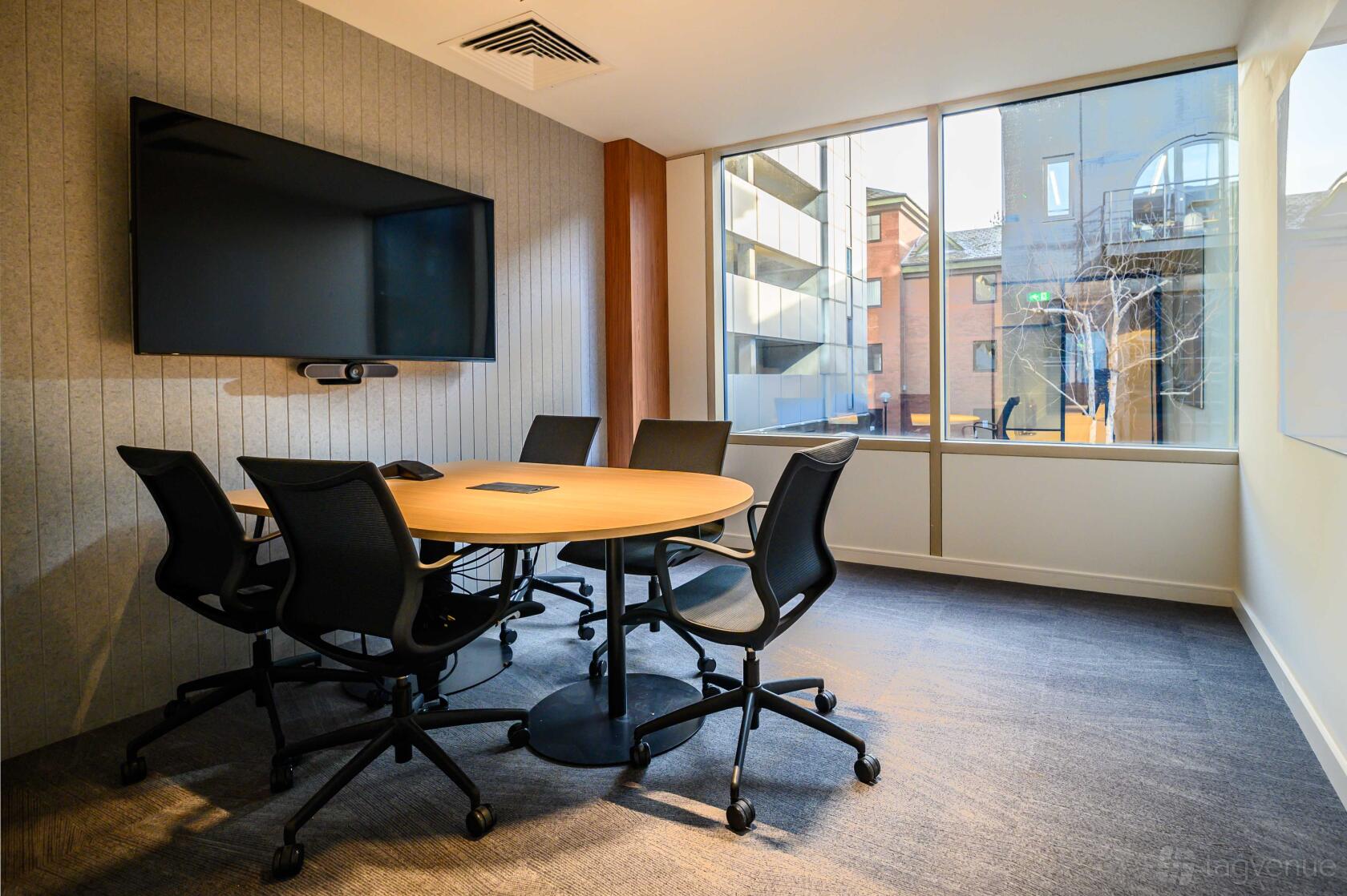 A boardroom with a round wooden table, black chairs, wall-mounted screen, and floor-to-ceiling windows at Serendipity Labs Manchester.