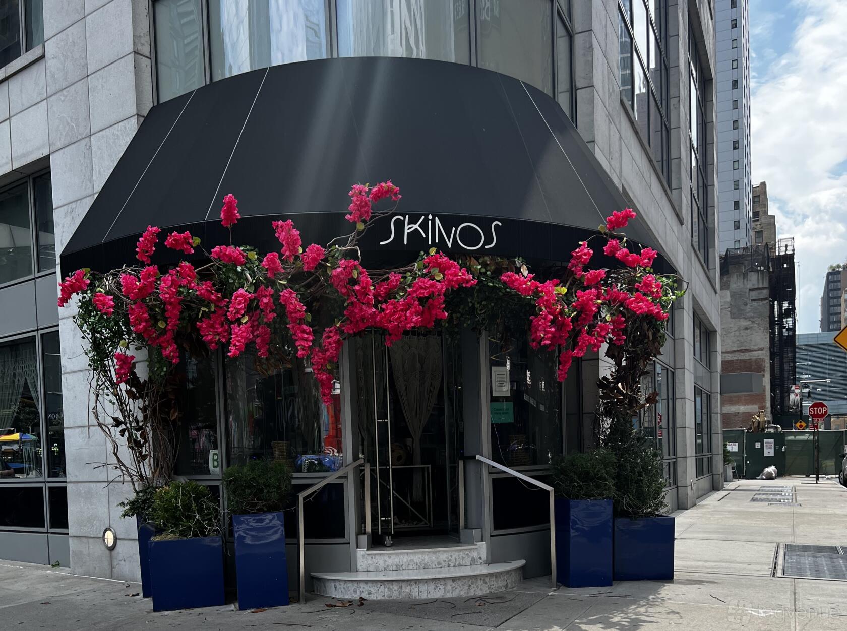 A restaurant entrance with black awning, large windows, and pink floral decorations at Skinos.