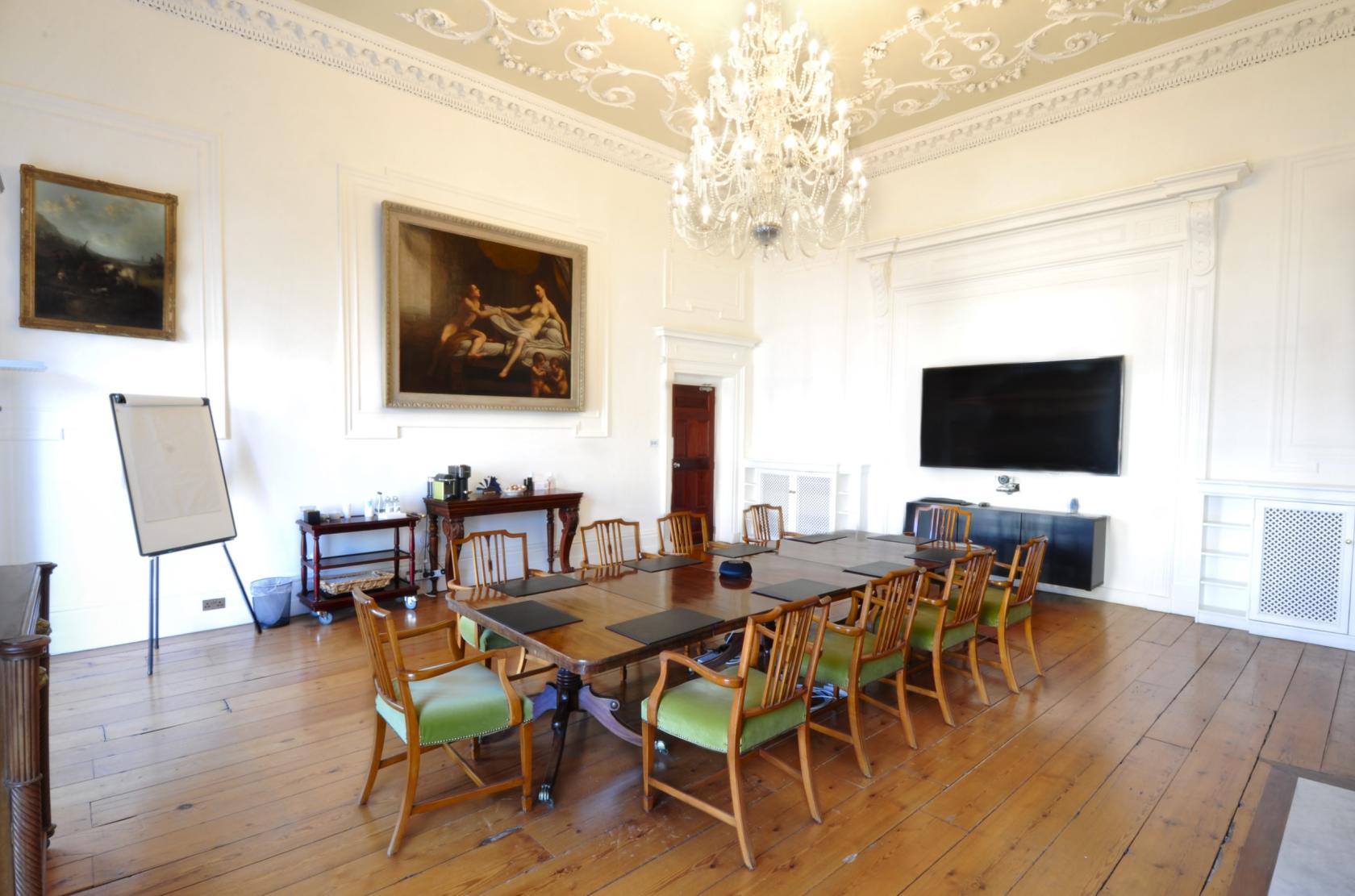 A boardroom with a long wooden table, green cushioned chairs, chandelier, and wall-mounted screen at 12 Merrion Square.