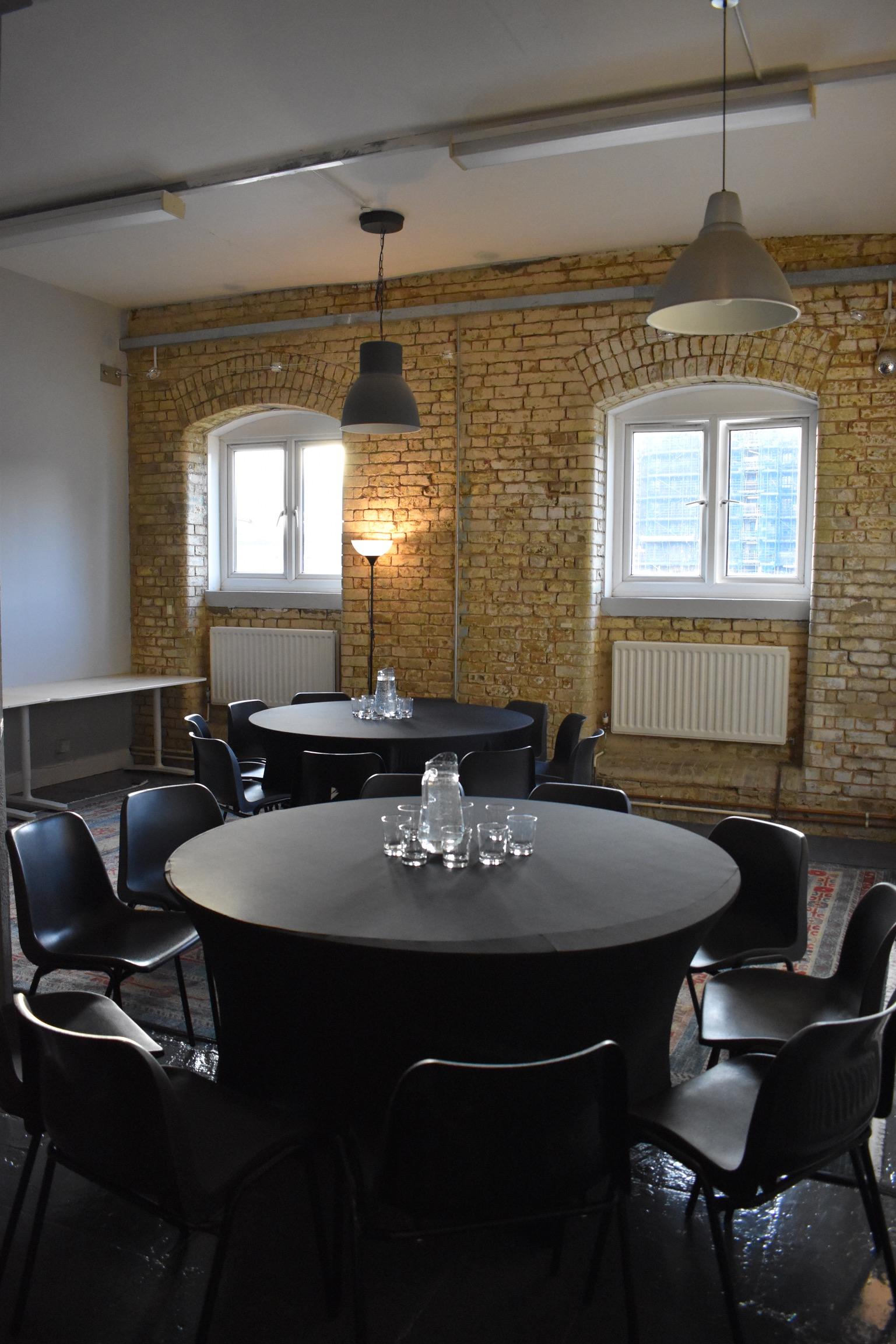 A function room with exposed brick walls, arched windows, and round tables set with water pitchers at Boathouse Studios, Barking.