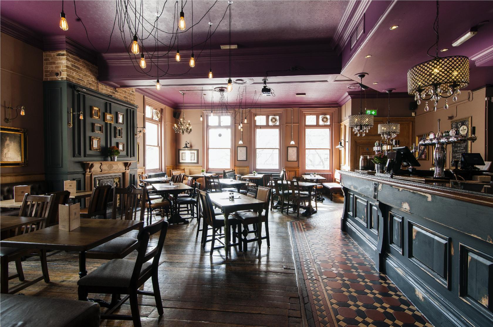 A function room in a pub with vintage wooden tables, chandeliers, exposed brick, and patterned tile flooring at The Marquis Cornwallis.