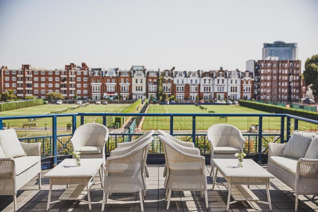 A function room terrace with white wicker seating, glass tables, and views of tennis courts at The Queen's Club.