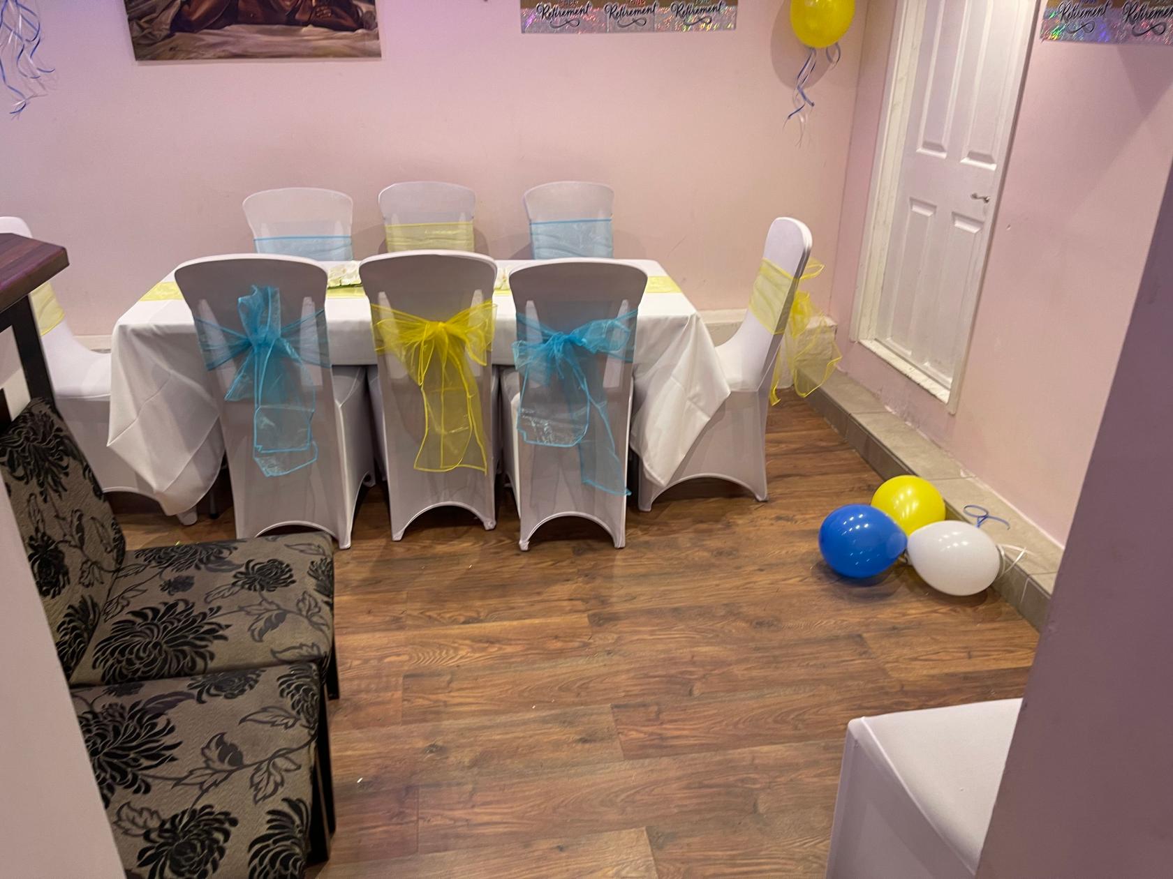 An event space with a long table covered in white linens, chairs draped with colorful bows, and balloons on wood flooring at Calabash Restaurant & Bar.