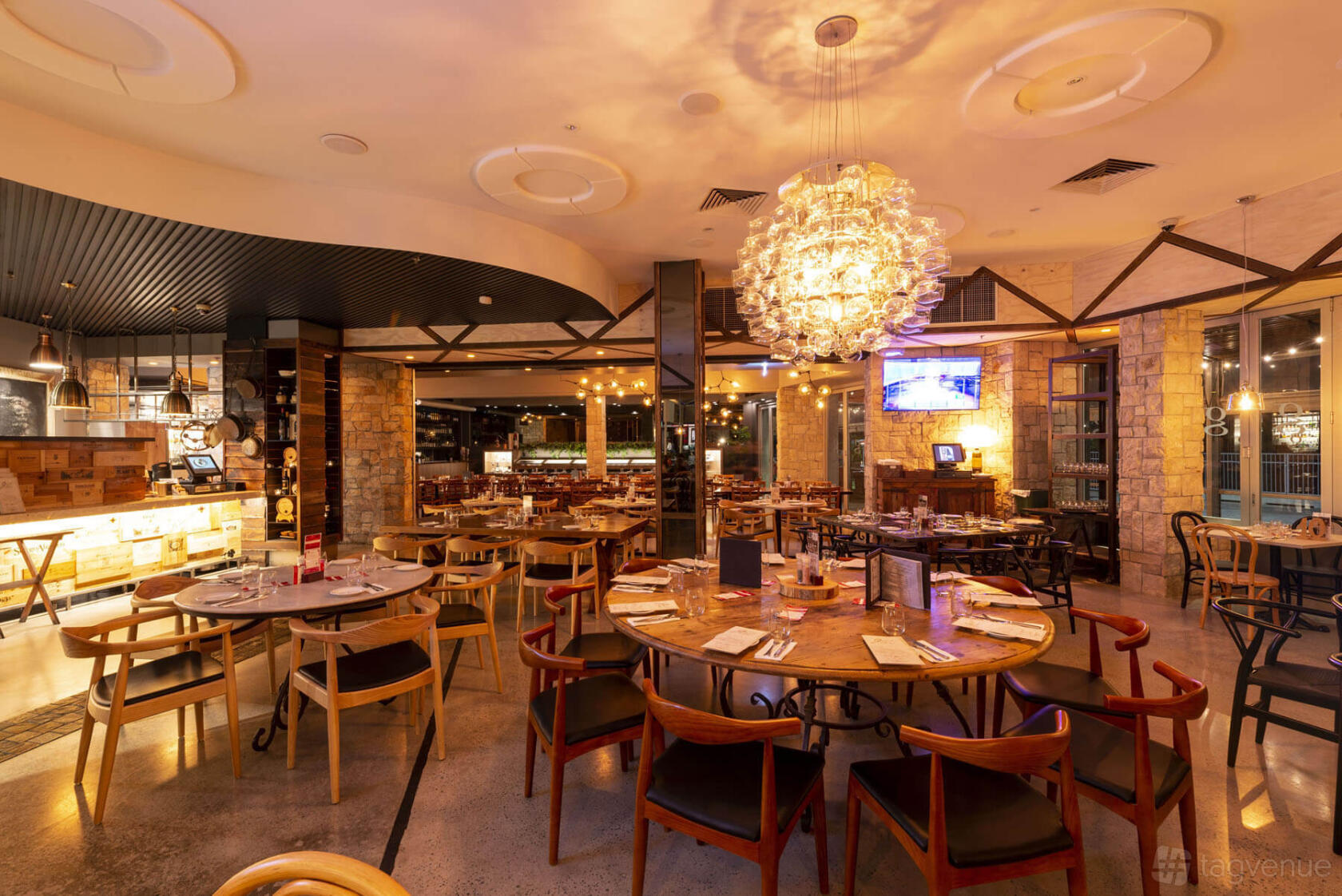 A restaurant dining area with round wooden tables, stone walls, and a large modern chandelier at Gusto da Gianni.