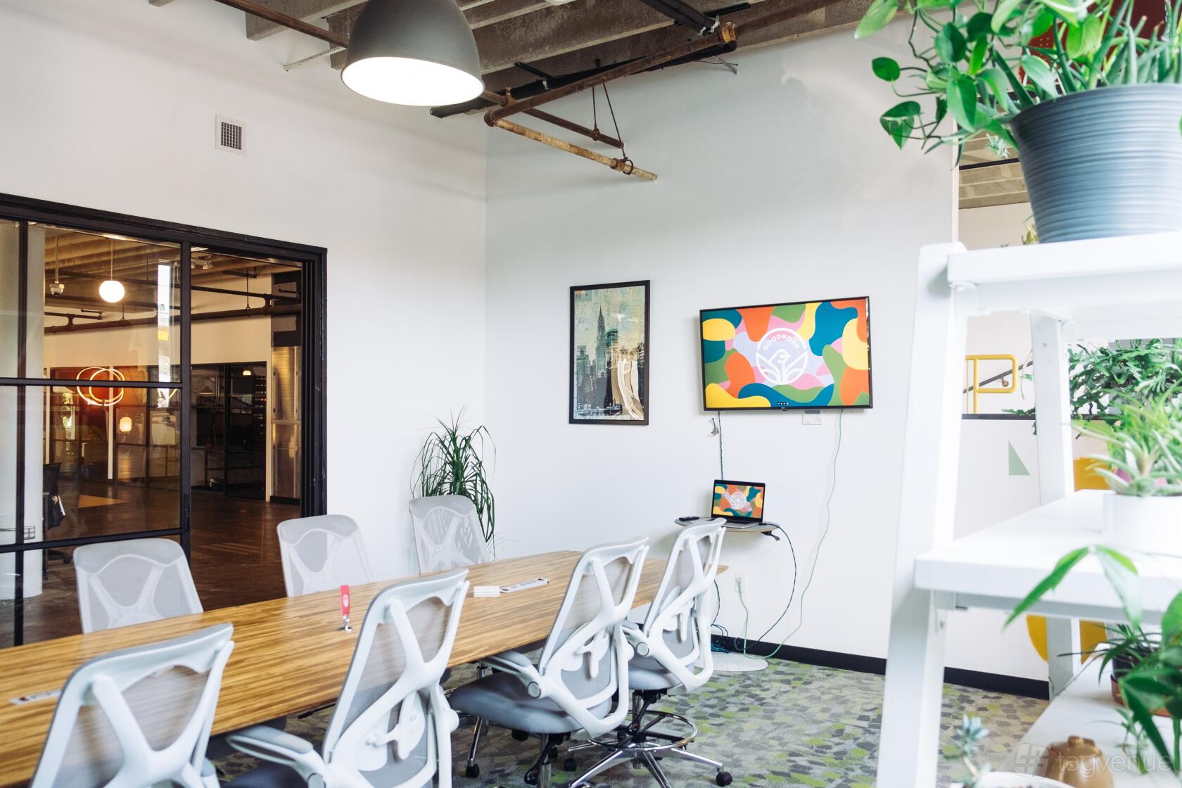 A meeting room with a long wooden table, white mesh chairs, wall-mounted TV, and potted plants at Good Coworking.