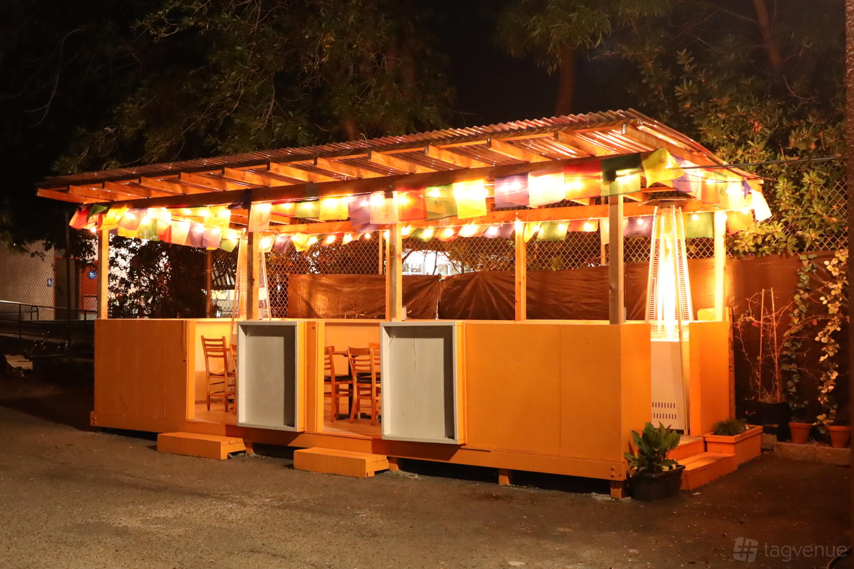 An outdoor restaurant patio with colorful string lights, wooden chairs, and patio heaters at Tashi Delek Party Palace.